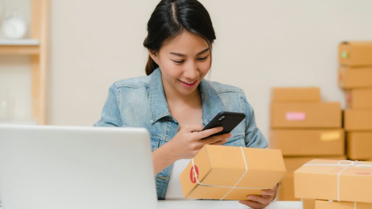 Person prüft ein Paket mit dem Handy neben Laptop und Kartons